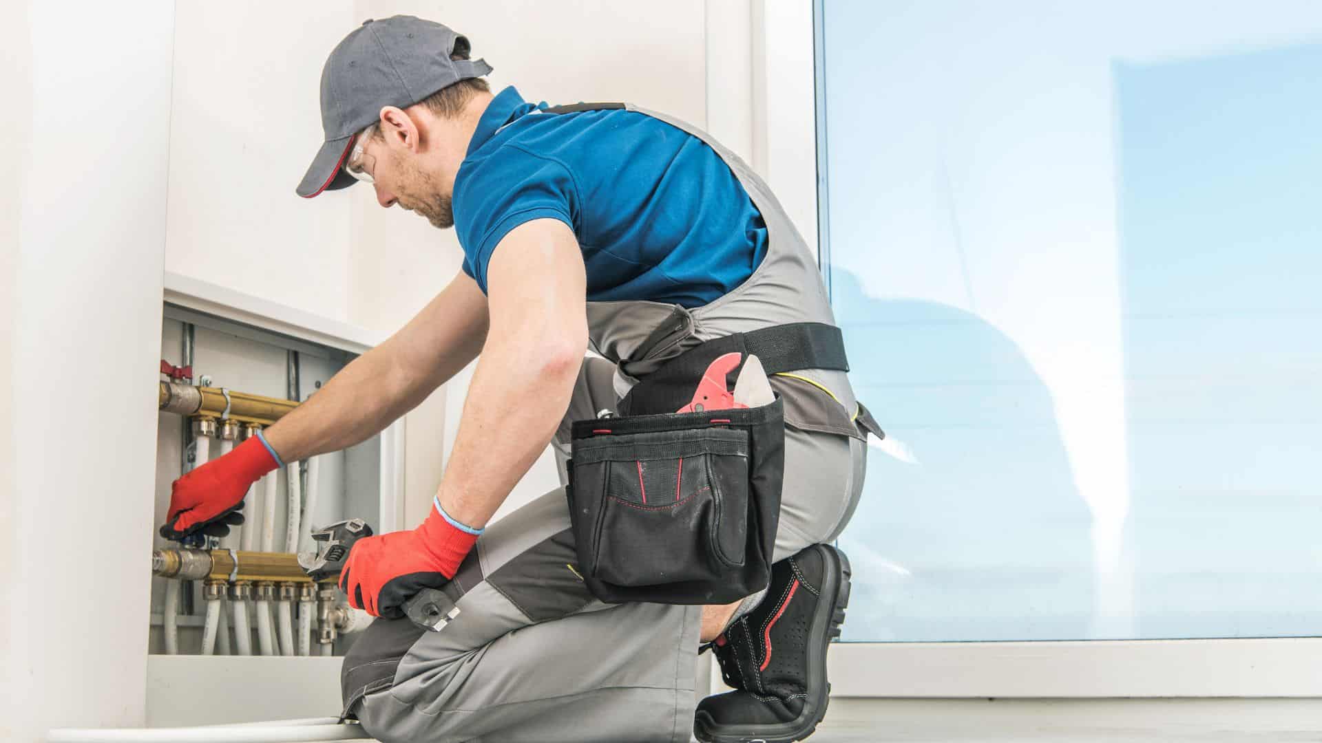 A plumber in a blue shirt and gray overalls is kneeling and working on pipes inside a building. He's wearing a cap, red gloves, and holds tools from his belt pouch. A large window is in the background.