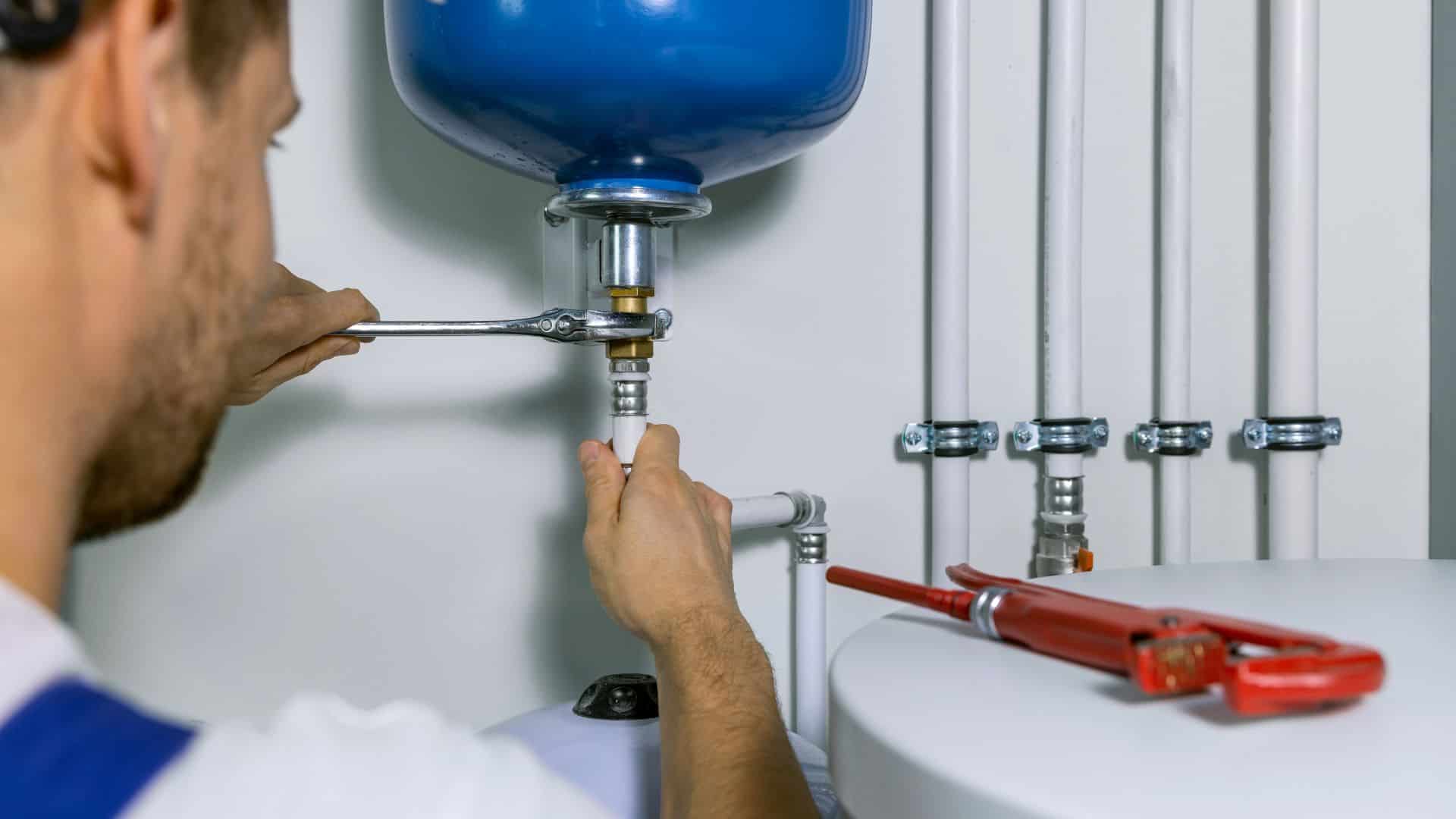 A plumber works on a large blue water tank, using a wrench to adjust a pipe connection. Several other pipes and valves are visible, with a red pipe wrench resting nearby. The setting is a utility room with white walls.
