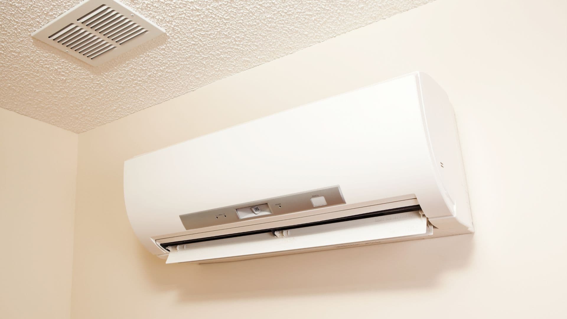 A wall-mounted air conditioning unit installed near the ceiling of a room, next to a ventilation grate. The unit is white with a curved design, blending into the cream-colored walls.