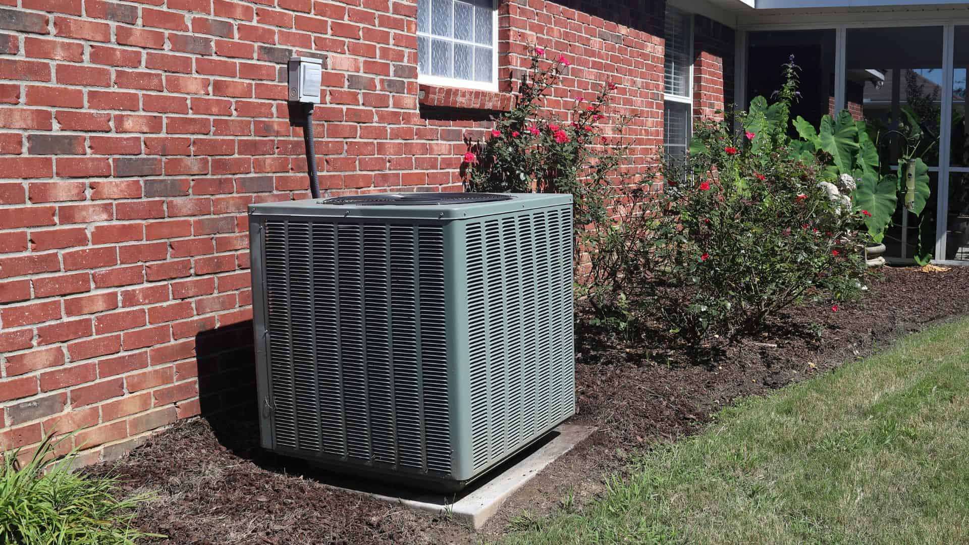 An outdoor air conditioning unit is positioned beside a red brick house. It's surrounded by well-maintained flower beds with green plants and blooming flowers. The area is in full sunlight, and the lawn is neatly trimmed.