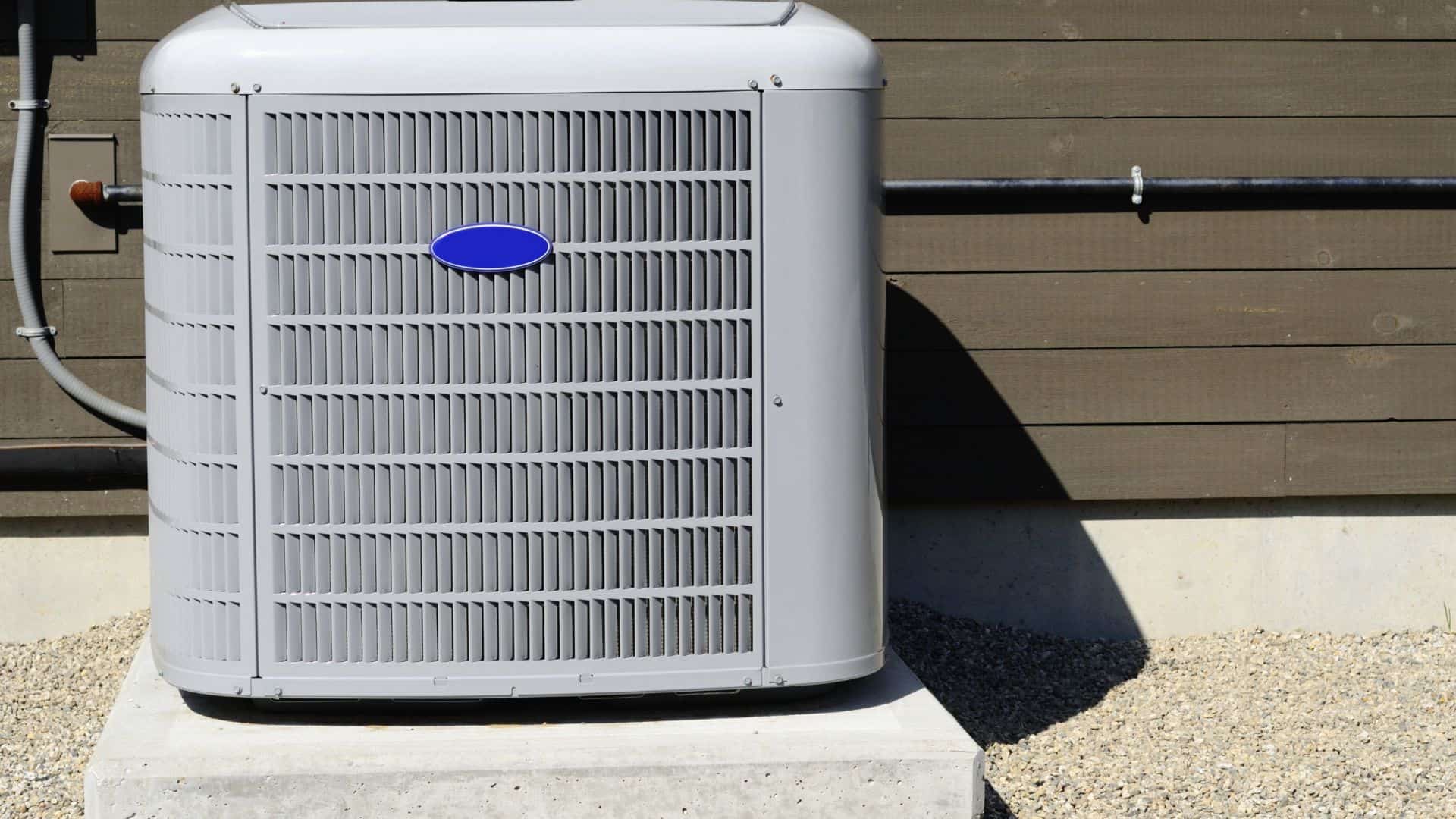 A large, gray outdoor air conditioning unit sits on a concrete slab. The unit is next to a wooden building with horizontal panels. Pebbles cover the ground surrounding the concrete base.
