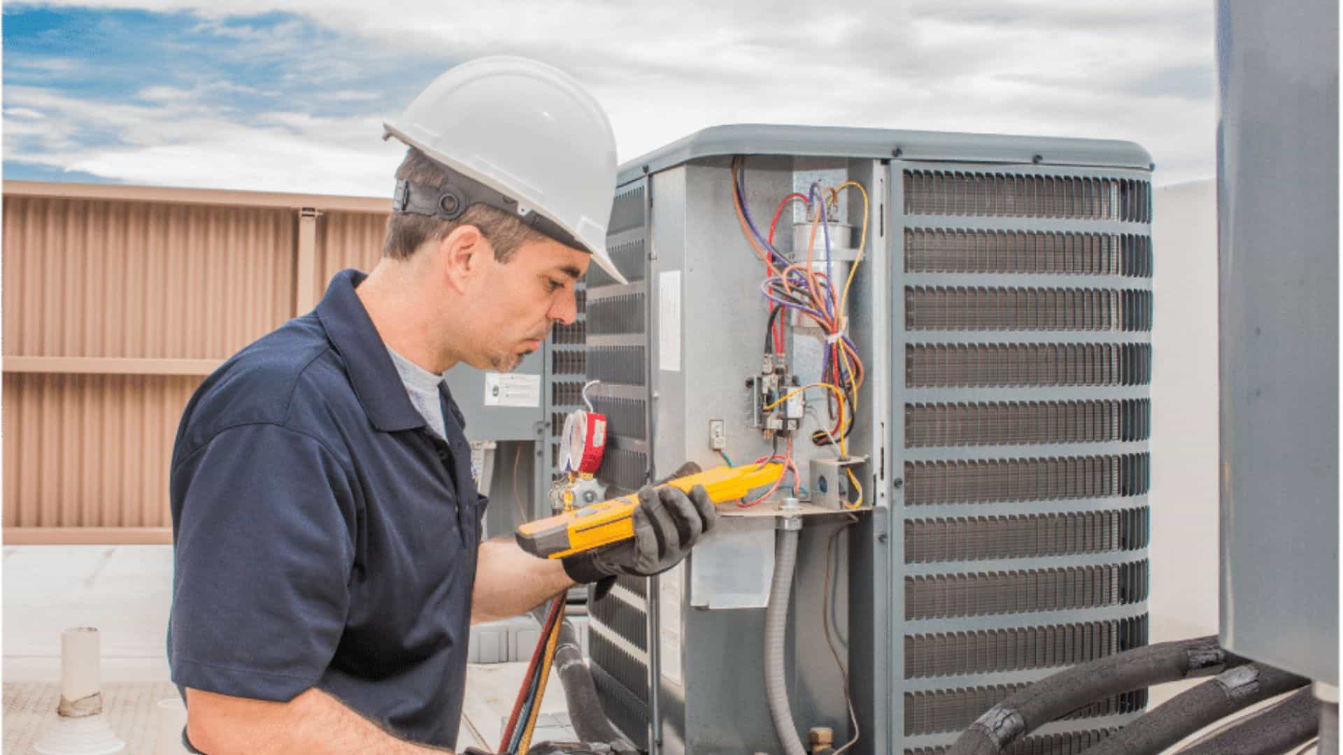 A technician in a hard hat and blue shirt uses a digital multimeter to inspect an open air conditioning unit on a rooftop. The sky is partly cloudy, and tools are visible on his utility belt.