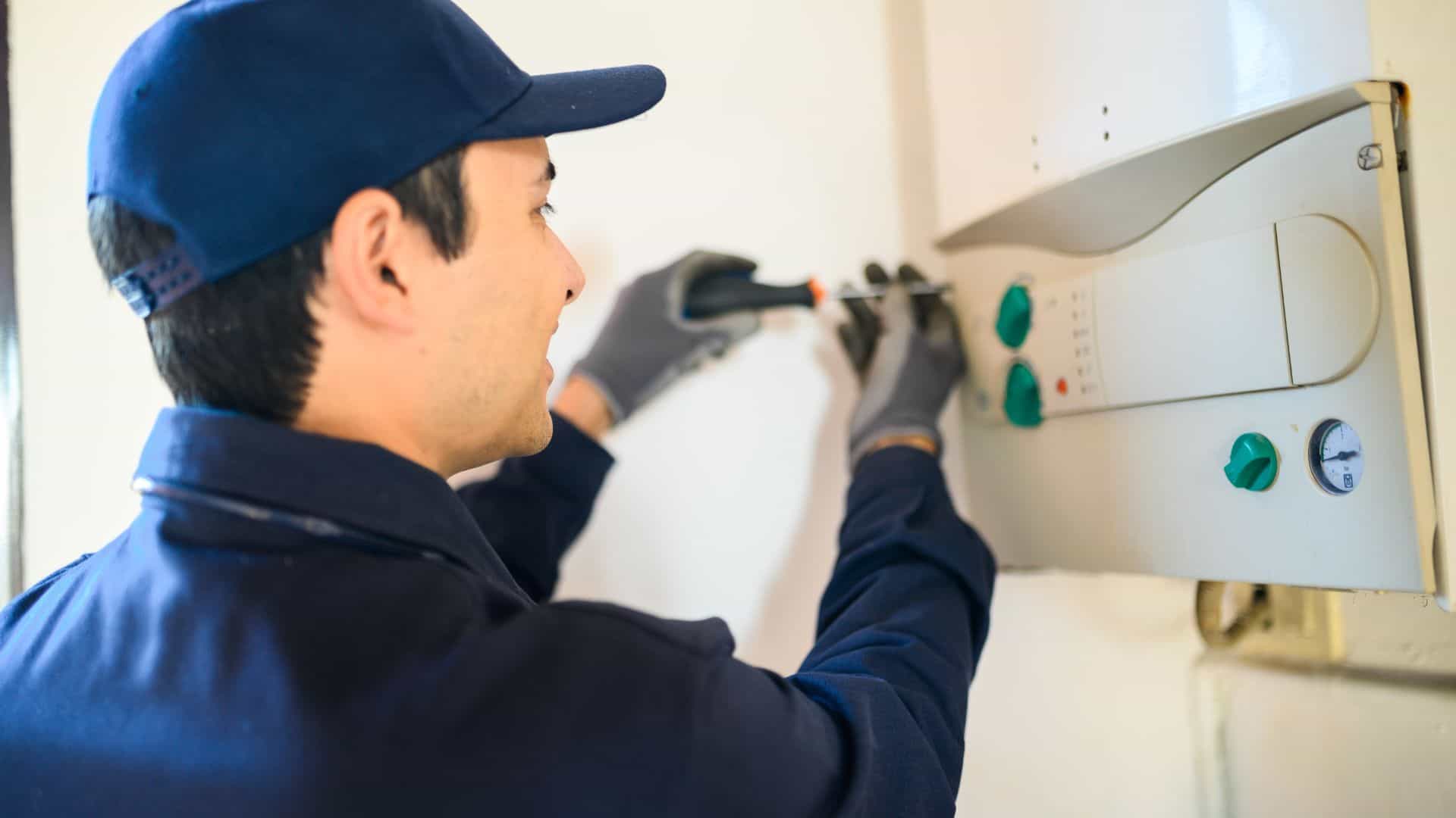 A technician wearing a blue uniform and cap is using a screwdriver to fix a control panel on a white wall. He is wearing gray gloves and focused on the task. The panel has various knobs and dials.