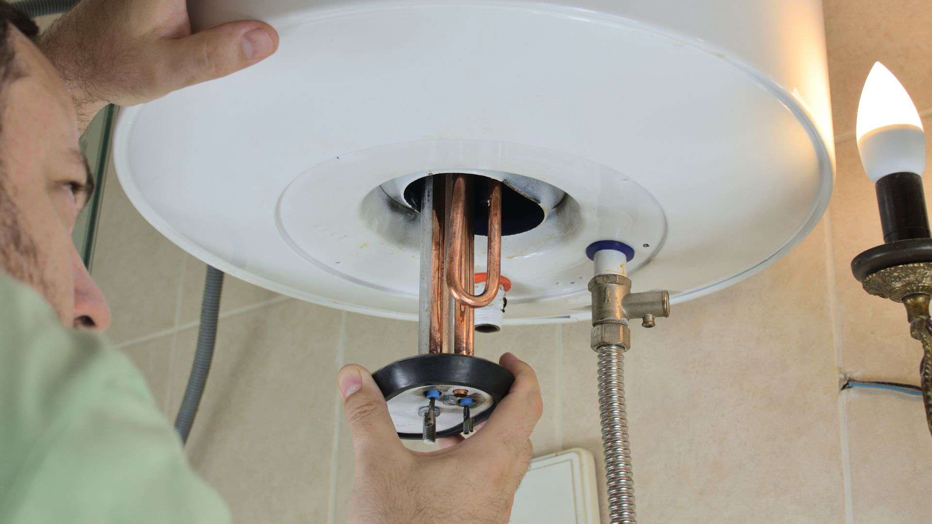 A person installing or repairing a water heater, focusing on the heating element. The image shows hands working with copper pipes and a metal fitting, alongside a lit candle, providing additional light. The background reveals tiled walls and a flexible water hose.