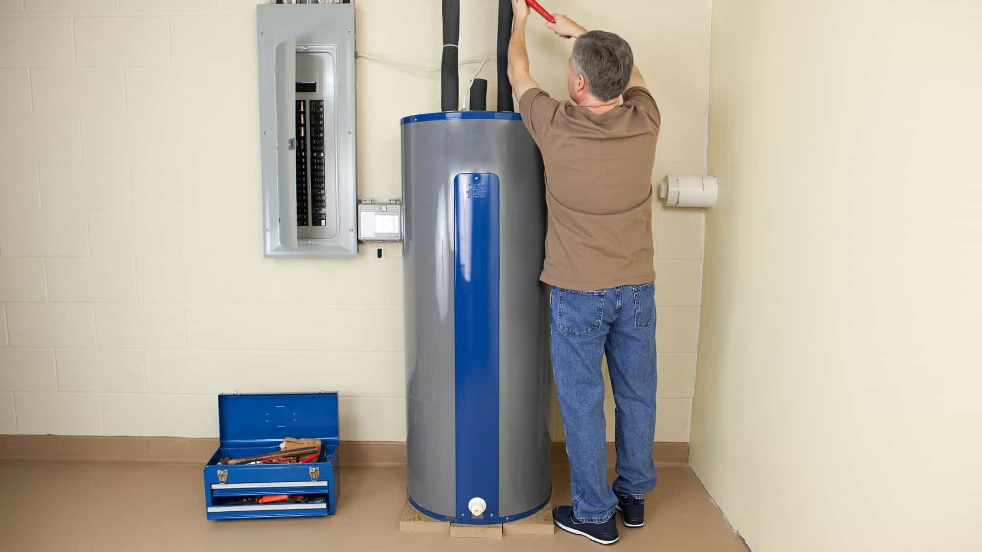 A person standing on a step stool is repairing a water heater in a utility room. There's an open blue toolbox with tools on the ground, and an electrical panel is visible on the wall.