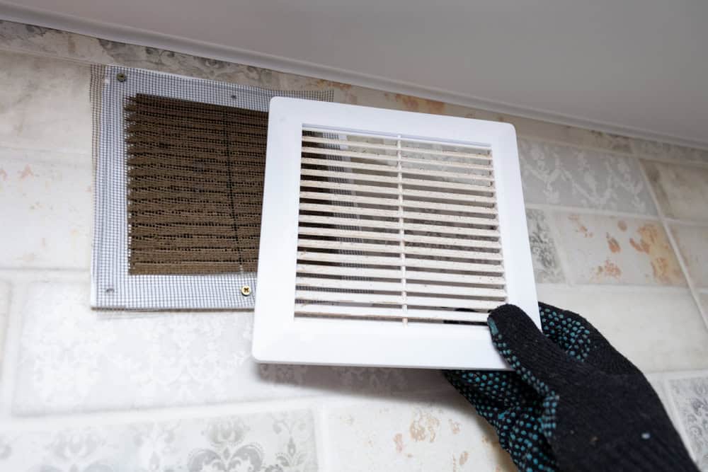 A gloved hand removing a dusty air vent cover from a wall, revealing a dirty filter behind it. The wall has a patterned tile design.