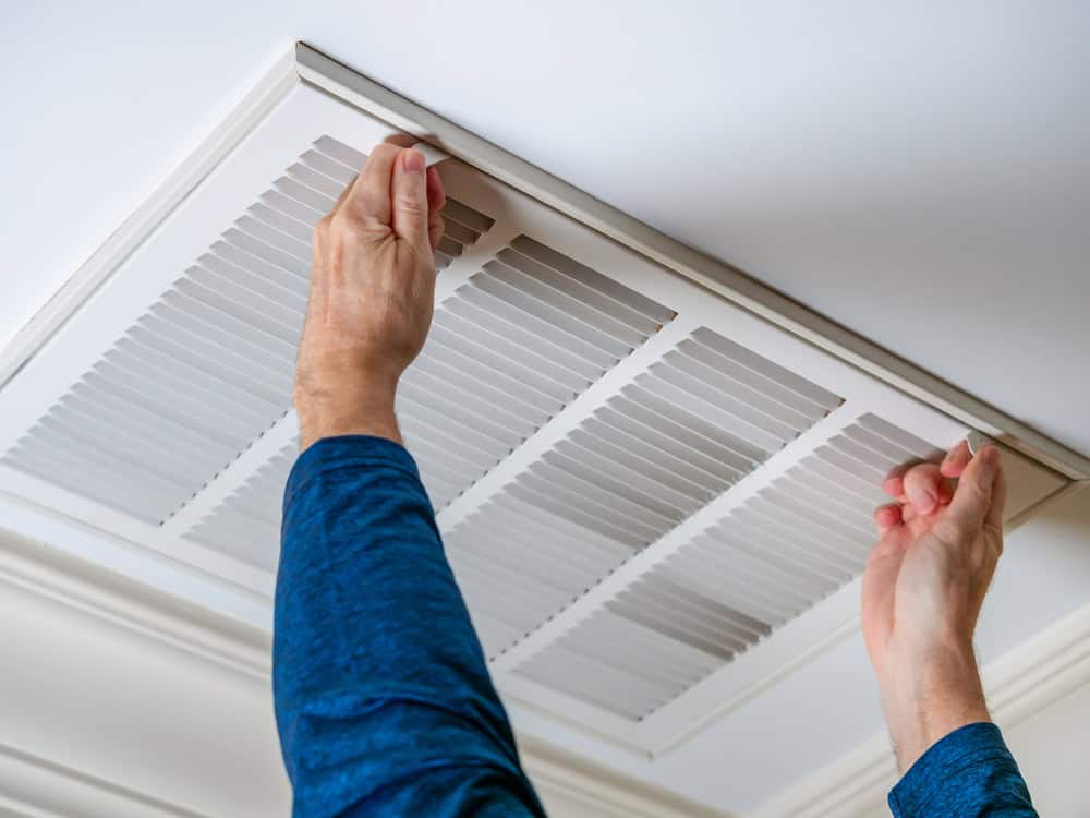 A person wearing a blue long-sleeve shirt is replacing or adjusting an air vent cover on a ceiling. The vent has a grid design, and the hands are positioned to secure it in place.