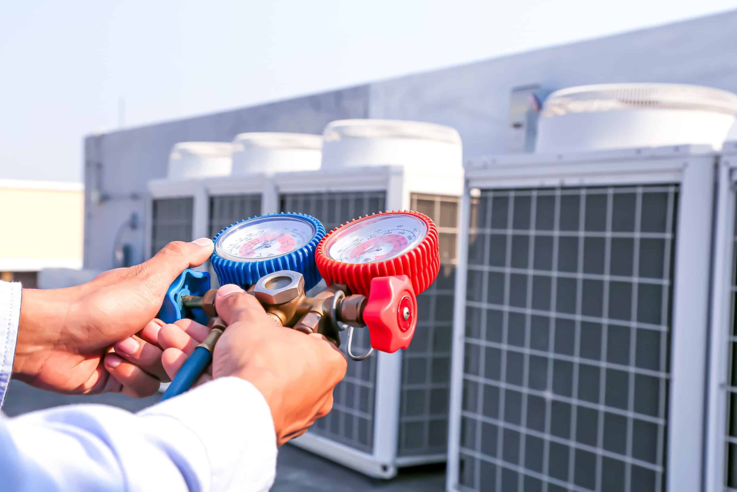 A technician holds a blue and red manifold gauge set in front of large outdoor air conditioning units on a sunny day, illustrating HVAC maintenance or repair work.
