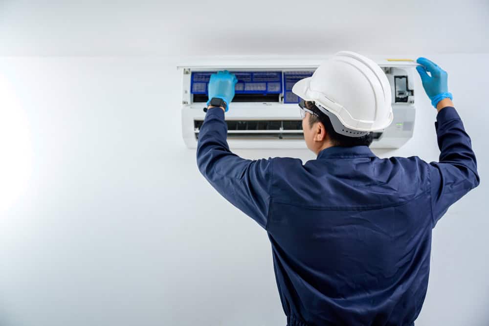 A person in a hard hat and gloves services an air conditioning unit mounted on a wall. They are adjusting the front panel while standing at the unit. The scene suggests maintenance or repair work in progress.