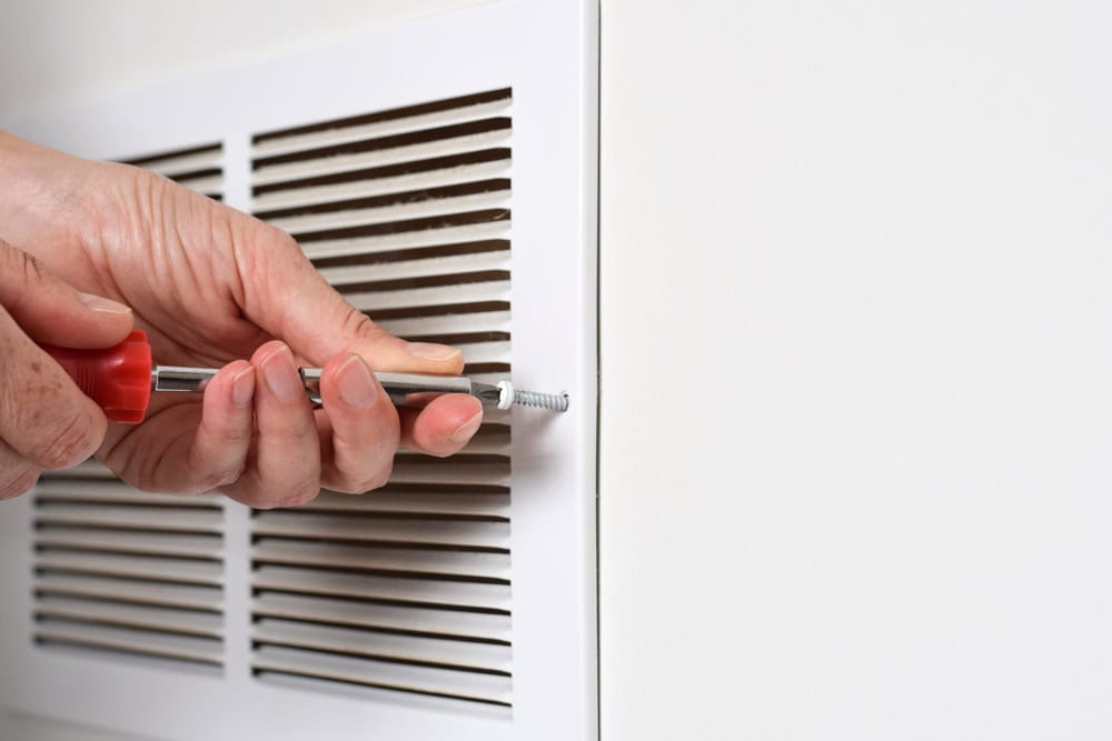 A person uses a screwdriver to tighten a screw on a white air vent cover attached to a wall. The focus is on the hands and the screwdriver.