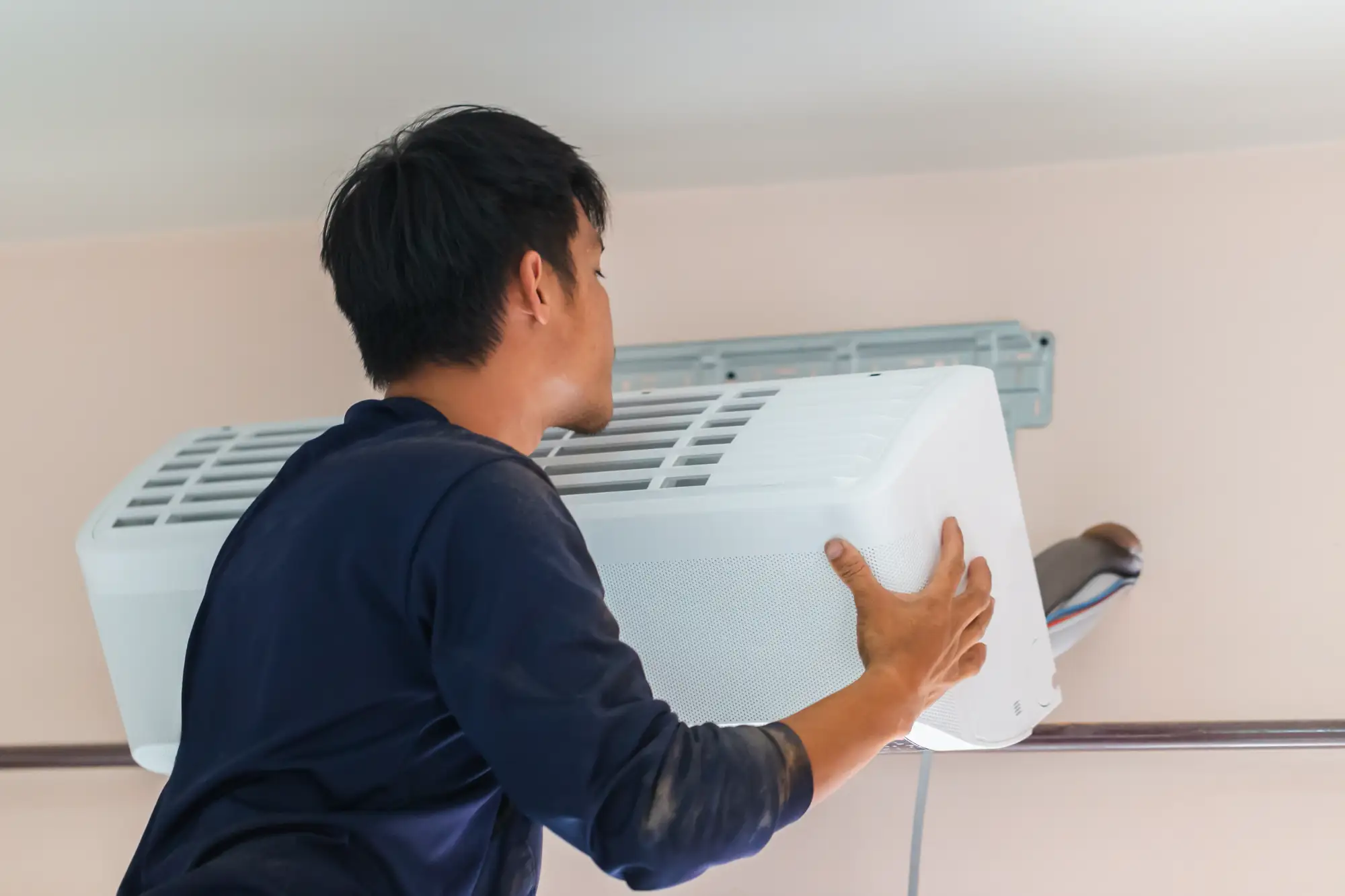 A technician wearing a dark shirt installs or repairs an air conditioner unit mounted high on a light-colored wall.