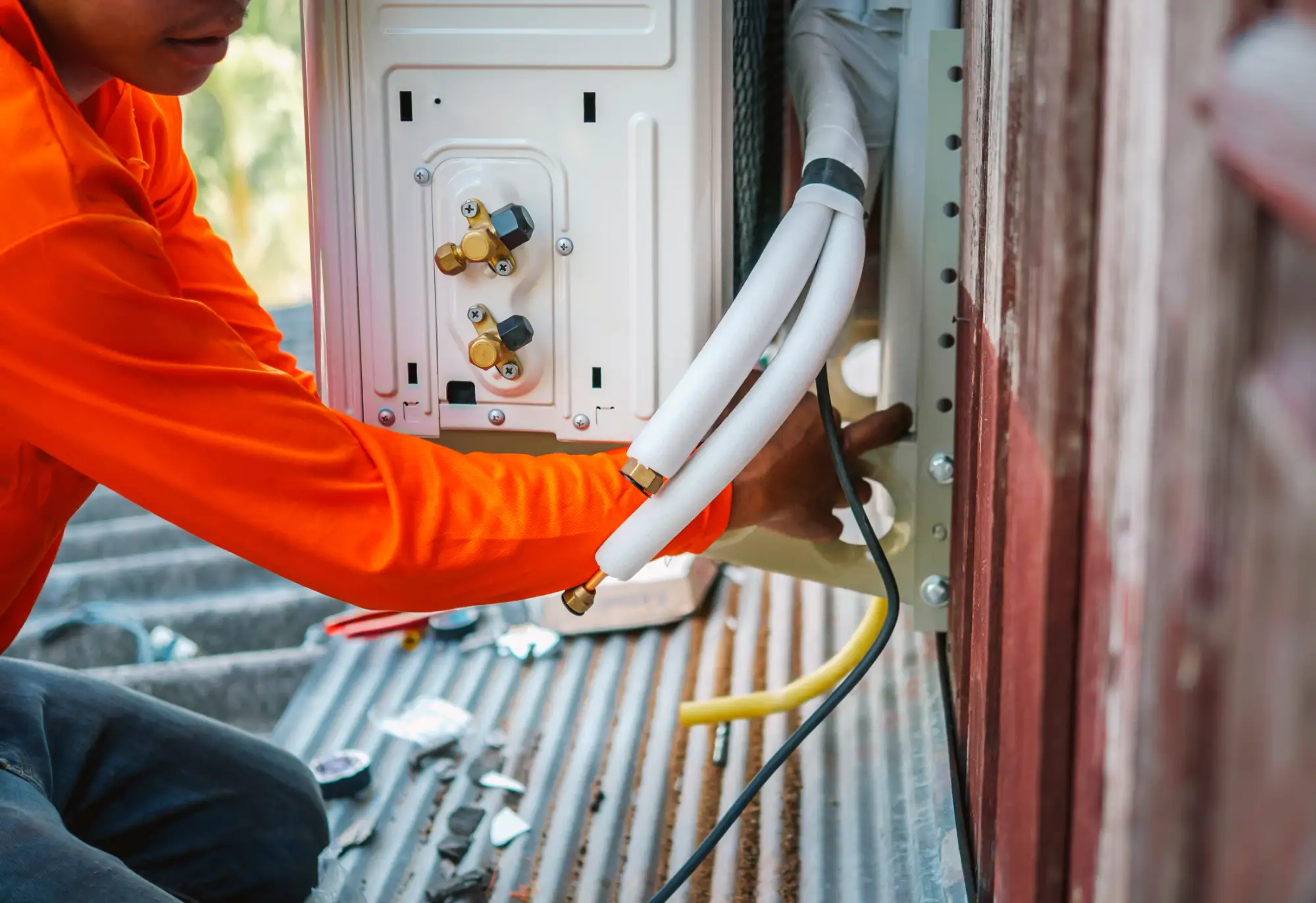 A person in an orange shirt installs or repairs an air conditioning unit on an exterior wall, adjusting pipes and connections while kneeling on a metal surface. Tools and equipment are scattered nearby.