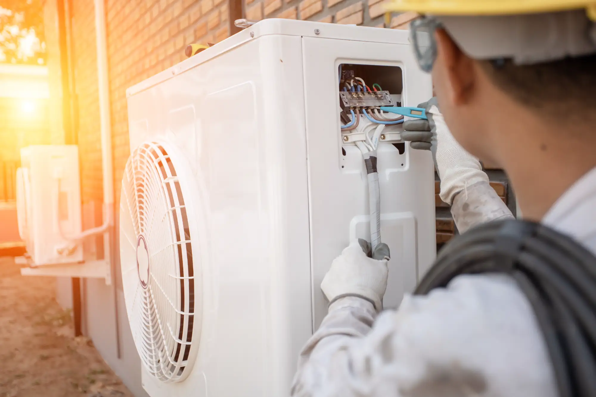 A technician wearing gloves and a hard hat repairs or installs an outdoor air conditioning unit using tools, with wiring visible inside the unit. The scene is lit by sunlight near a brick wall.