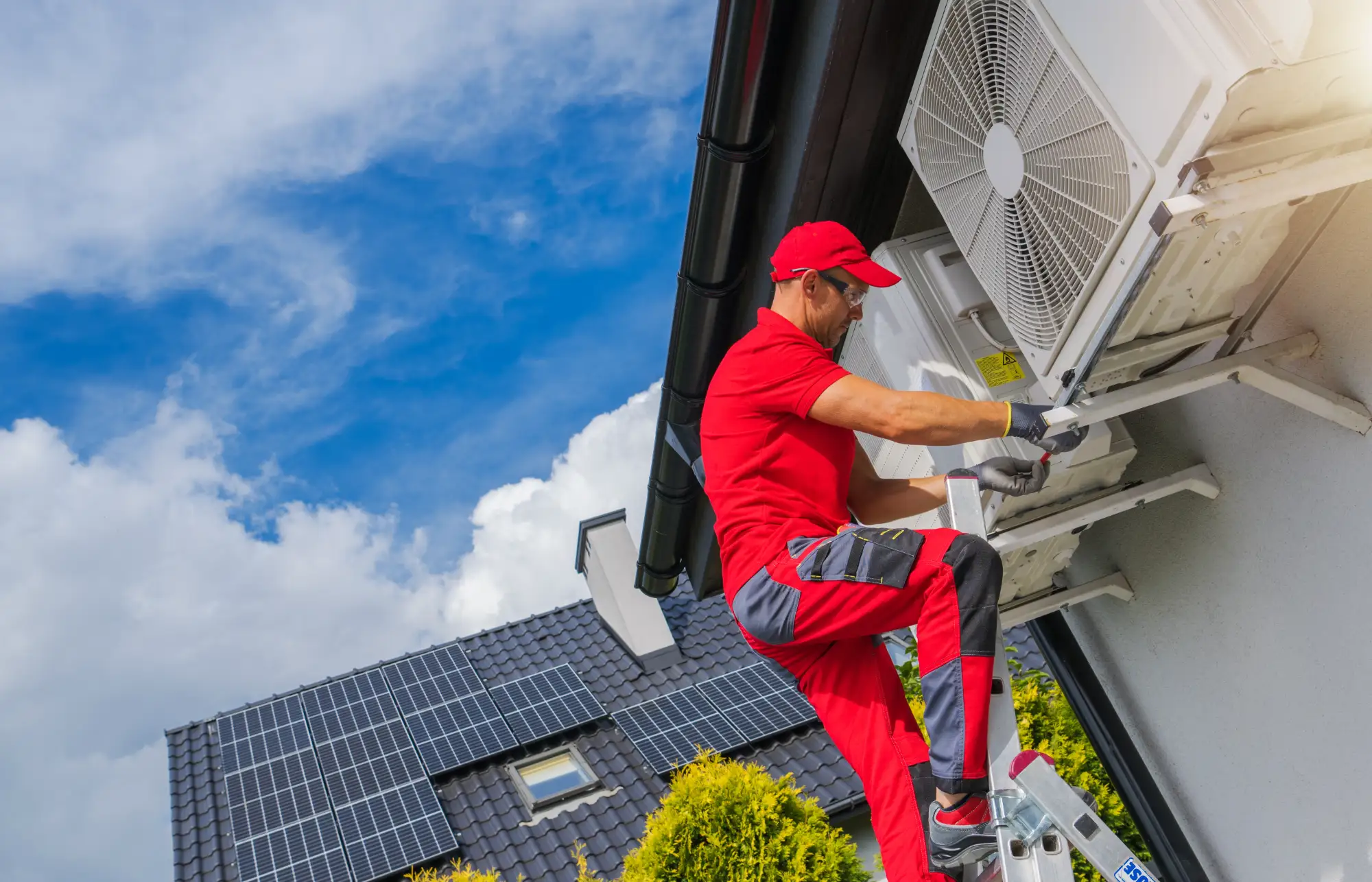 A technician in red uniform stands on a ladder installing or servicing an outdoor air conditioning unit on a house with solar panels on the roof, with a blue sky and clouds in the background.