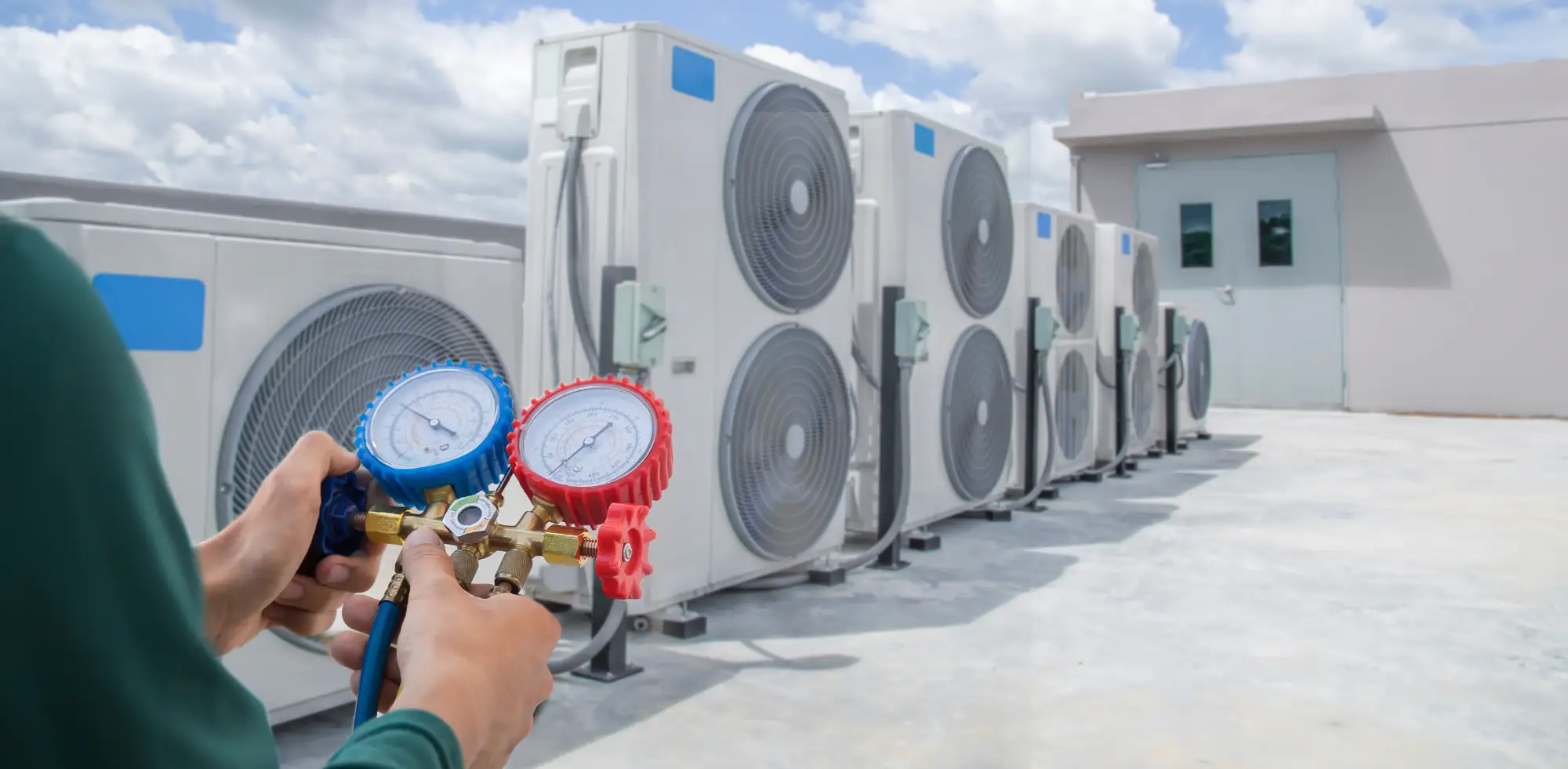 A person holds HVAC pressure gauges in front of a row of outdoor air conditioning units on a rooftop under a partly cloudy sky.