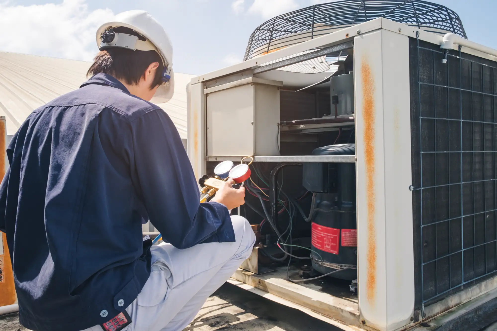 A technician wearing a helmet and safety glasses uses gauges to inspect or repair an outdoor air conditioning unit on a rooftop under a partly cloudy sky.
