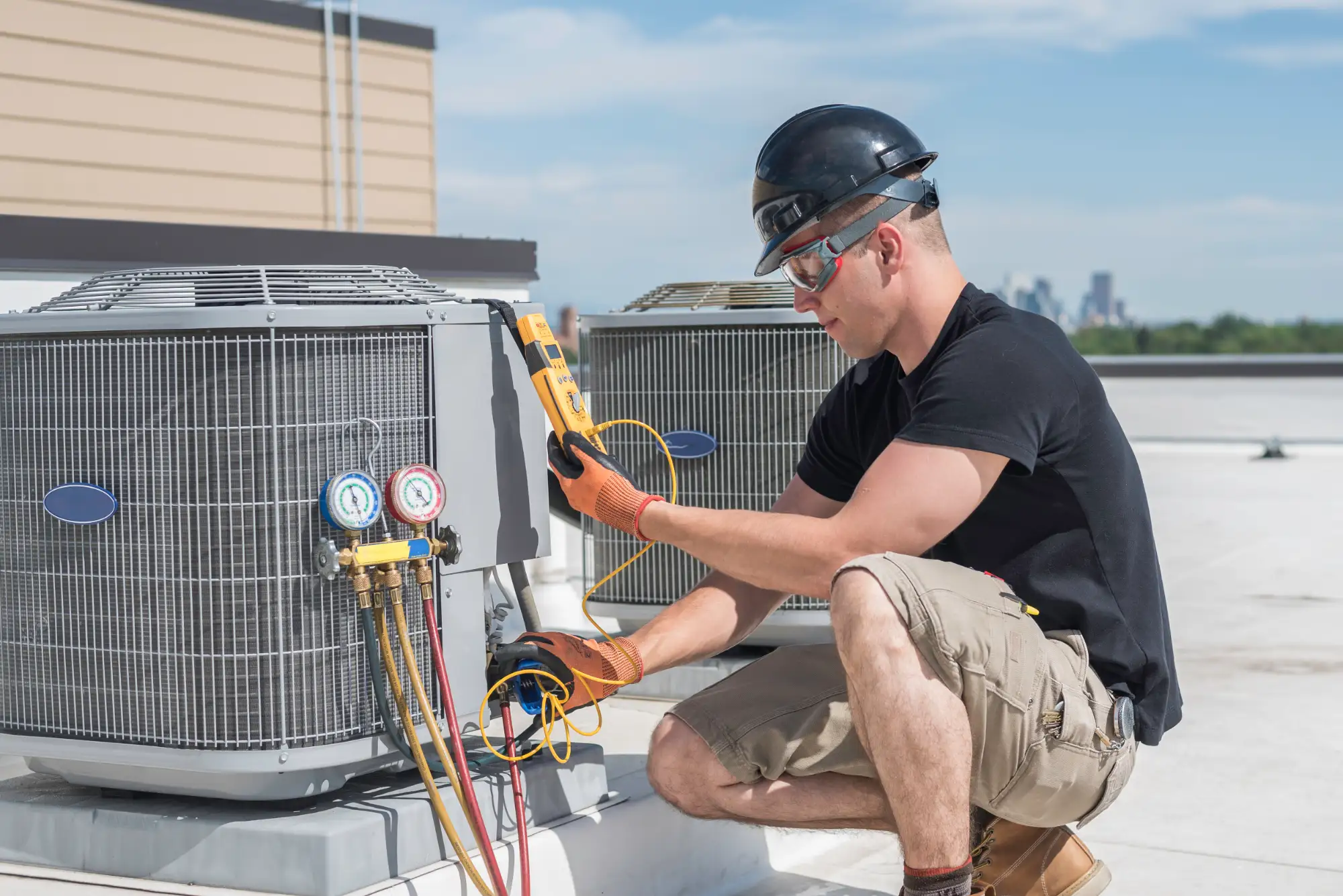 A technician wearing safety gear kneels on a rooftop and tests a commercial air conditioning unit with a digital multimeter on a sunny day.