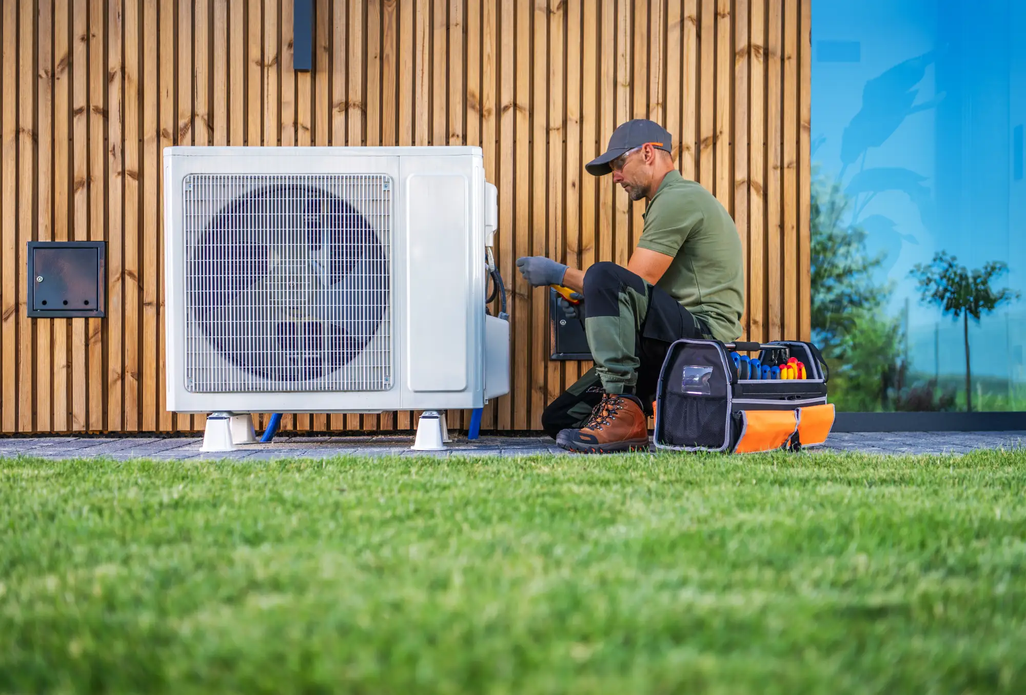A technician in work clothes kneels beside an outdoor air conditioning unit, using tools from a kit to perform maintenance or repairs, with a modern wooden building and grass lawn in the background.
