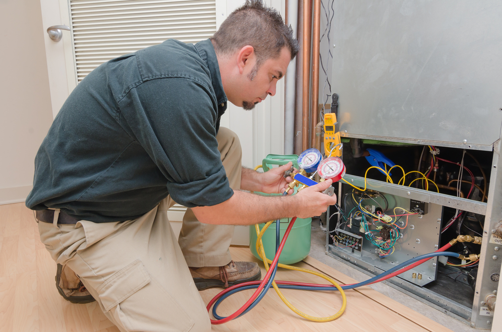 A technician kneels on the floor, using gauges and tools to inspect or repair an HVAC unit with its panel open, surrounded by hoses and wires.