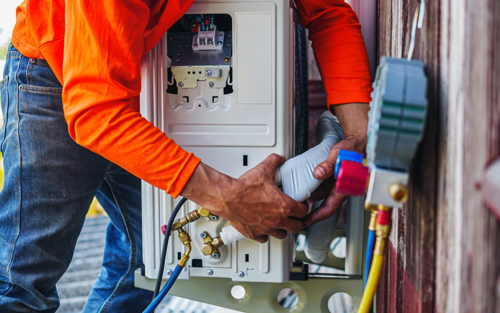 A technician in an orange shirt installs or repairs an air conditioning unit mounted on a wall, adjusting pipes and wires with gauges and tools visible nearby.